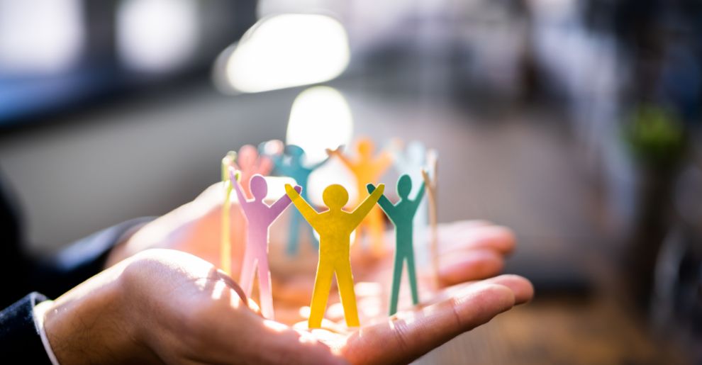 A close-up of two open hands gently cupping a circle of colourful paper cut-out figures, each with their arms raised, symbolising unity and support. The figures are in a variety of colours (yellow, orange, blue, green, and purple), standing in a ring formation. The background is softly blurred, with warm natural light creating a calm, inclusive atmosphere. This image conveys themes of community, collaboration, and care.